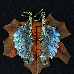 May include: Earrings made from iridescent abalone shell, featuring brass-coloured metal detailing. The earrings are designed as stylised feathers, showcasing blue, green, and purple hues. They are presented on a brown, leaf-shaped fabric.