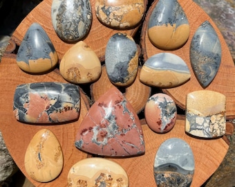 May include: A collection of polished gemstones in various shapes, including ovals, teardrops, and rectangles. The stones display a range of colors, such as beige, gray, and reddish-brown, with unique patterns and textures. They are arranged on a wooden surface.