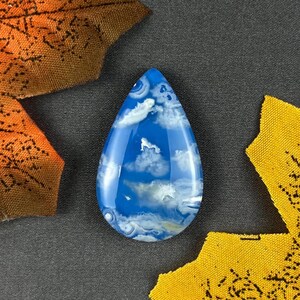Könnte beinhalten: Ein tropfenförmiger Edelstein mit einem Muster aus blauem Himmel und Wolken. Der polierte Stein hat eine glänzende Oberfläche mit weißen und hellblauen Wirbeln, die an Wolken erinnern. Der Edelstein liegt vor einem dunkelgrauen Hintergrund mit gelben und orangen Blattakzenten.