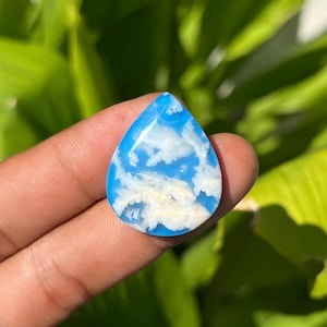 Könnte beinhalten: Ein tropfenförmiger Edelstein mit einer leuchtend himmelblauen Grundfarbe und komplizierten, wolkenartigen weißen und blassgelben Einschlüssen auf der Oberfläche. Der Stein hat eine glatte, polierte Oberfläche und eine markante Spitze.