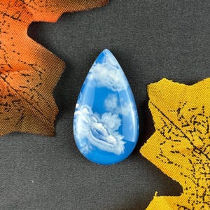 Pode incluir: Um pingente em forma de gota com um design de céu azul e nuvens. O pingente é azul brilhante com redemoinhos brancos que se assemelham a nuvens. O pingente é cercado por folhas de outono laranja e amarelas em um fundo cinza escuro.