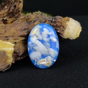 May include: An oval gemstone with a blue and white cloud design. The polished stone is set against a dark background with a piece of wood. The gemstone's design mimics a daytime sky with fluffy clouds.