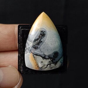 Könnte beinhalten: Ein dreieckiger, polierter Achat-Cabochon mit einem einzigartigen Muster aus Gelb, Weiß, Grau und Schwarz. Der Stein hat eine glatte, glänzende Oberfläche und ist in einer schwarzen Präsentationsbox eingefasst.
