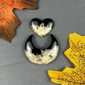 Puede incluir: Dos colgantes de resina: un corazón blanco y negro y una forma de media luna blanca y negra. Los colgantes tienen un acabado brillante y un patrón arremolinado único. El fondo es de color gris sólido, con acentos de hojas naranjas y amarillas.