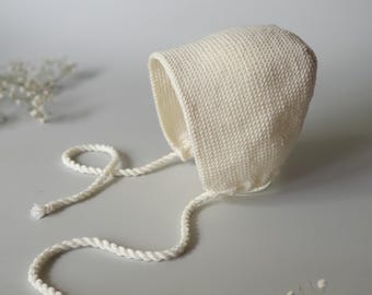 May include: A white crocheted baby bonnet with a white rope tie. The bonnet is laying on a white surface.