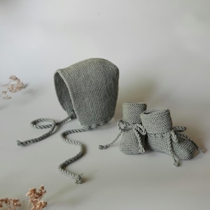 Peut inclure: Ensemble pour bébé en tricot gris comprenant un bonnet à liens et une paire de chaussons assortis. Le bonnet a une forme arrondie et un bord festonné. Les chaussons ont un revers et sont noués avec du fil assorti.