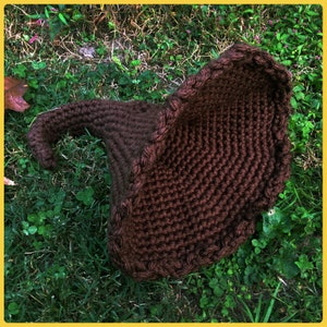 May include: A brown crocheted witch's hat with a pointed tip and a wide brim. The hat is lying on a bed of green grass.