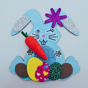 Puede incluir: Una manualidad de conejo azul con purpurina, sosteniendo una zanahoria y rodeado de huevos de Pascua. El conejo tiene una nariz rosa y un ojo que guiña.