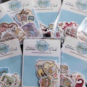 May include: A collection of sticker packs in clear plastic bags. Each pack has a light blue backing card with the words "Sticker Prints Pack" and the Etsy shop's URL. The stickers feature various colorful illustrations.