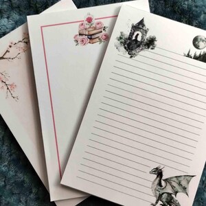 May include: Three white notepads with different designs. One features a cherry blossom branch, another has roses and books, and the third has a dragon, castle, and moon illustration. Each notepad has lined pages.