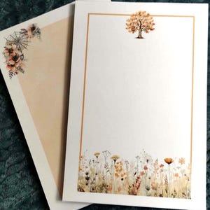 May include: Two rectangular sheets of stationery paper. The top sheet features a thin gold border, a small watercolor tree at the top center, and a field of wildflowers at the bottom. The sheet underneath shows a spiderweb and floral corner design.