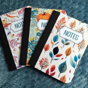 May include: Three rectangular notebooks with black spines and white covers featuring colourful autumn-themed patterns of leaves, flowers, and pumpkins. Each notebook has a central white label with a decorative border and the word NOTES printed in teal.