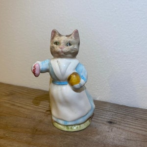 Puede incluir: Una figurita de cerámica de un gato con un vestido azul y blanco y sosteniendo una pelota amarilla. El gato tiene pelaje marrón y blanco y ojos verdes.