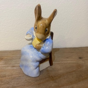 May include: A ceramic figurine of a brown rabbit wearing a blue dress and a yellow scarf, sitting on a wooden chair and holding a cup.