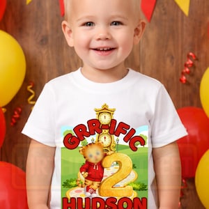 May include: A smiling toddler wearing a white t-shirt with a colorful graphic. The graphic features a cartoon tiger, the word "GRIFFIC", the number "2", and the name "HUDSON". The design is set against a green landscape with a clock.