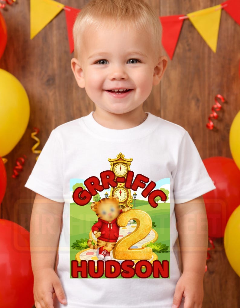 May include: A smiling toddler wearing a white t-shirt with a colorful graphic. The graphic features a cartoon tiger, the word "GRIFFIC", the number "2", and the name "HUDSON". The design is set against a green landscape with a clock.