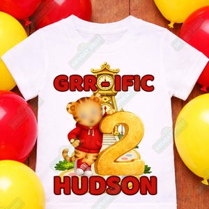May include: White t-shirt with a cartoon tiger wearing a red hoodie and holding a large gold number 2. The shirt has the text "GRROIFIC" and "HUDSON" in red. Red and yellow balloons are in the background.