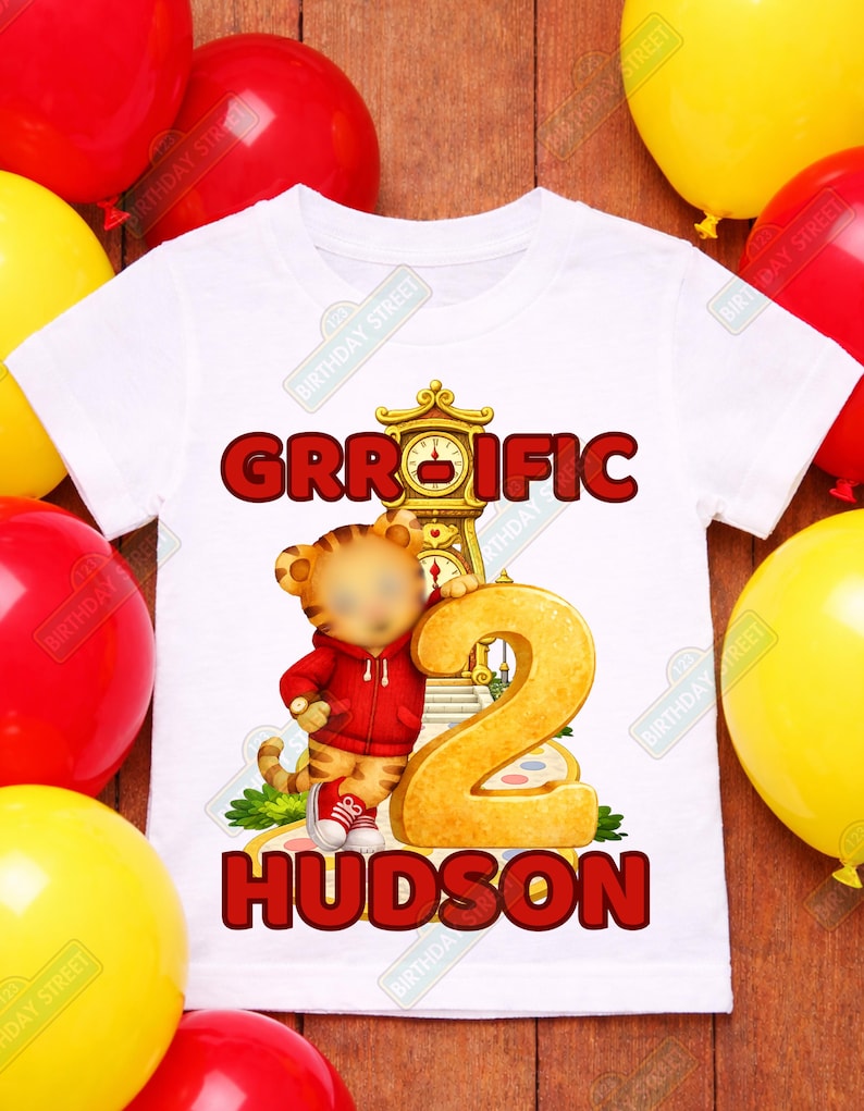 May include: White t-shirt with a cartoon tiger wearing a red hoodie and holding a large gold number 2. The shirt has the text "GRROIFIC" and "HUDSON" in red. Red and yellow balloons are in the background.