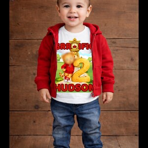 May include: A young child wearing a red hooded jacket, blue jeans, and red sneakers. The child is smiling and wearing a white t-shirt with a cartoon graphic and the name "HUDSON". The graphic includes the number "2".