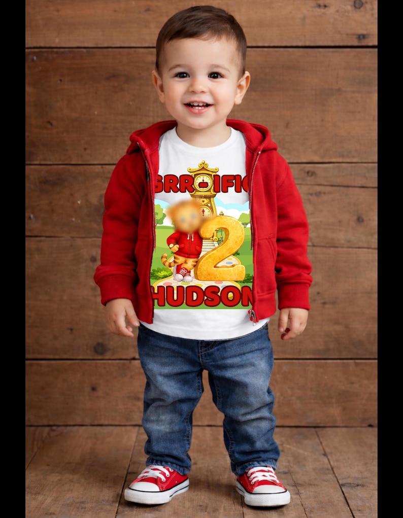 May include: A young child wearing a red hooded jacket, blue jeans, and red sneakers. The child is smiling and wearing a white t-shirt with a cartoon graphic and the name "HUDSON". The graphic includes the number "2".