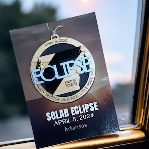 May include: A wooden ornament shaped like the state of Arkansas with the word "ECLIPSE" in blue letters. The ornament is decorated with a gold star and the text "Path of Totality" and "RUSTY TRACTOR VINEYARDS-LITTLE ROCK, AR". The ornament is labeled "SOLAR ECLIPSE APRIL 8, 2024 Arkansas".
