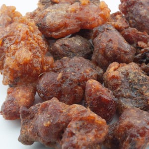 May include: Close-up of a pile of dried, amber-colored resin pieces. The pieces vary in size and shape, with a slightly translucent appearance. The resin has a glossy sheen, suggesting a natural product. The background is plain white.