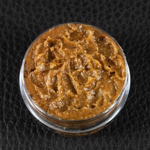 Peut inclure: Un petit pot transparent rempli d'une substance brune texturée. Le contenu semble épais et légèrement huileux, avec une couleur riche et terreuse. Le pot est posé sur un fond sombre et texturé, mettant en valeur l'apparence du produit.