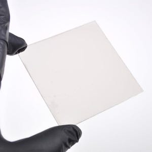 Op de afbeelding: Een vierkant, doorschijnend, gebroken wit vel wordt vastgehouden door een gehandschoende hand. Het vel lijkt gemaakt te zijn van een dun, flexibel materiaal. De achtergrond is effen wit, wat een helder contrast met het object oplevert.