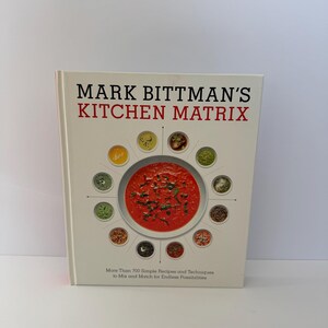 May include: A hardcover cookbook titled "MARK BITTMAN'S KITCHEN MATRIX" with a white cover. The cover features a circular design of various food items surrounding a bowl of red soup. The text below reads "More Than 700 Simple Recipes and Techniques to Mix and Match for Endless Possibilities."