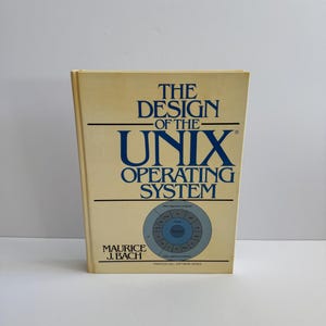 以下が含まれることがあります： モーリス・J・バッハ著「The Design of the UNIX Operating System」というタイトルのハードカバー本。本はベージュ色の表紙で、青と黒の文字と円形の図があります。Prentice Hall Software Seriesのロゴが下にあります。