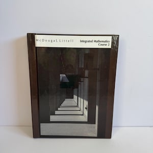 Puede incluir: Un libro de texto titulado "McDougal, Littell Integrated Mathematics Course 2". La portada presenta una imagen arquitectónica oscura de un largo corredor con pilares y sombras repetidas. El libro tiene una lomo marrón.