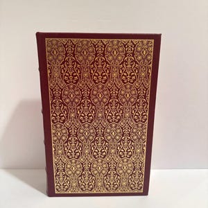 May include: A burgundy book with a gold, ornate pattern on the cover. The pattern is a repeating design of swirling shapes and floral elements. The book's spine is visible on the left side.