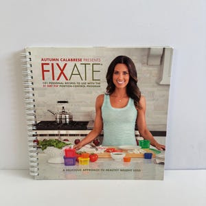 May include: A spiral-bound cookbook titled "FIXATE" by Autumn Calabrese. The cover features a woman in a light blue tank top. The book's title and subtitle are visible, along with the phrase "A Delicious Approach to Healthy Weight Loss."