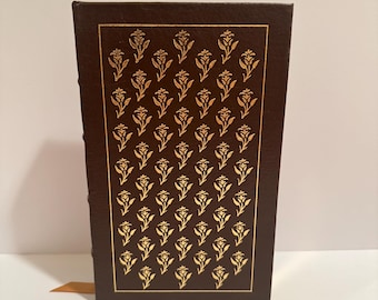 May include: A hardcover book with a dark brown cover and gold floral pattern. The book has a gold-colored border and a matching gold ribbon bookmark. The spine is also dark brown.