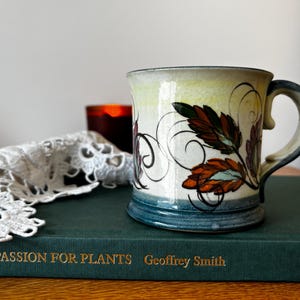 Può includere: Una tazza in ceramica dipinta a mano con un motivo a foglie nei toni dell'arancione, del marrone e del blu. La tazza ha una parte superiore color crema e una base e un manico blu. La tazza è su un libro verde con il testo "A Passion for Plants".