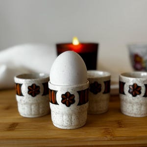 Può includere: Quattro portauova in ceramica maculata con un motivo floreale marrone e arancione. Una tazza contiene un uovo bianco. Le tazze sono su una superficie di legno, con una candela accesa e una tazza da tè sullo sfondo.