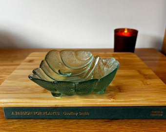 Op de afbeelding: Een doorschijnende groene glazen schaal in de vorm van een blad met een getextureerd oppervlak en geschulpte randen. De schaal heeft een verhoogd aderpatroon aan de binnenkant en rust op kleine glazen voetjes, met een helder ontwerp.