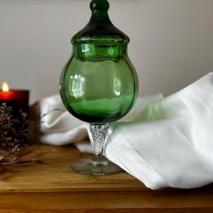 Puede incluir: Un tarro de boticario de vidrio verde con tapa, sobre una superficie de madera. Una servilleta de tela blanca está doblada cerca. Una vela encendida en un soporte rojo oscuro y flores secas también están sobre la mesa.