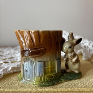 May include: A ceramic egg cup shaped like a brown tree stump with a sculpted door and window. A small, light brown rabbit figure is attached to the side of the stump, standing on a green base that mimics grass. The finish is glossy and smooth.
