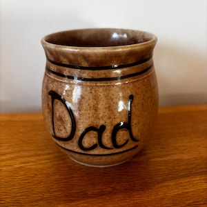 Puede incluir: Una taza de cerámica marrón con la palabra "Dad" en escritura negra. La taza tiene una textura moteada y dos rayas negras cerca de la parte superior. La taza está sobre una superficie de madera.
