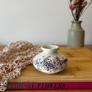May include: A small, round, cream-colored ceramic vase with a crackle glaze finish, featuring a hand-painted floral pattern in shades of pink, blue, and green. The vase has a short, narrow neck and a wide, bulbous body with a smooth, rounded base.