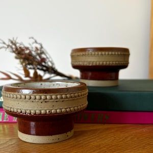 Peut inclure: Deux bols en céramique sur pied, présentant un vernis bicolore marron et beige. Le bord supérieur est orné d'une texture perlée en relief, tandis que la partie inférieure possède une finition marron lisse et brillante sur une base beige mouchetée.