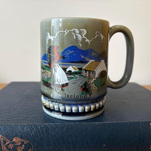 Może przedstawiać: Ceramiczny kubek z błyszczącym wykończeniem w kolorze oliwkowej zieleni, ozdobiony kolorową ilustracją irlandzkiego krajobrazu z postacią, białym domkiem, niebieskimi górami i napisem Ireland w kolorze czarnym. Podstawa posiada ozdobny koralikowy brzeg.