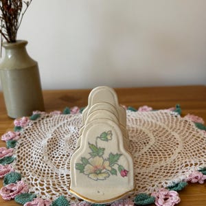 May include: A cream-colored ceramic toast rack featuring a delicate floral illustration of a yellow and pink flower with green leaves on the front panel. The rack has a curved, arched design with gold-toned trim along the edges of the dividers.