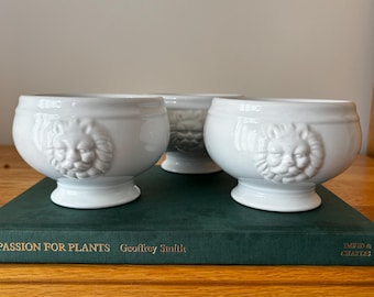 May include: Three white ceramic bowls, each featuring a raised, sculpted lion head design on the front. The bowls have a rounded body, a smooth glossy finish, and a pedestal base. They are arranged in a cluster with two in the front and one behind.