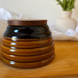 May include: A brown ceramic jar with a wooden lid. The jar has a black upper section and a ribbed design. The jar is on a wooden surface.