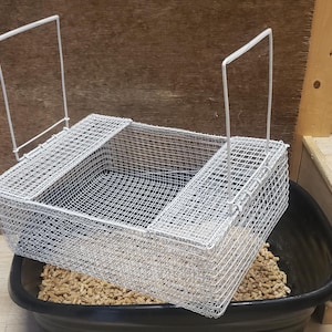 May include: A rectangular metal wire cage with a handle sits inside a black container filled with small, brown pellets. The cage has a mesh design and is angled. The cage is likely for small animals.