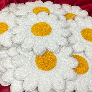 May include: A collection of white daisy-shaped decorations with yellow centres. The petals have a fluffy, textured appearance. The daisies are arranged on a red surface, providing a striking contrast. These items are likely for home decoration.