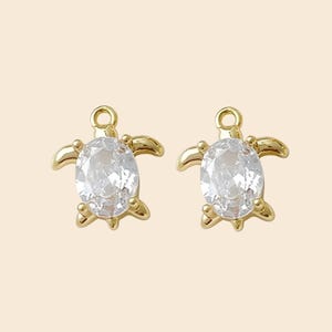 May include: Two gold-colored turtle charms, each featuring a clear, oval-shaped gemstone as the turtle's body. The charms have small loops at the top for attaching to jewelry. The background is a solid, light beige color.