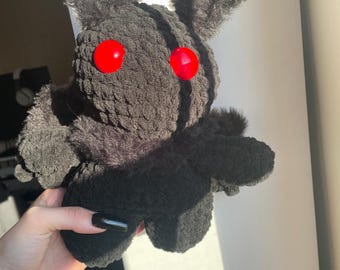 May include: A handmade black plush toy with large red button eyes and fluffy ears. The plush has a fuzzy texture and is held in a person's hand. The toy's design resembles a creature with a unique, slightly spooky aesthetic.